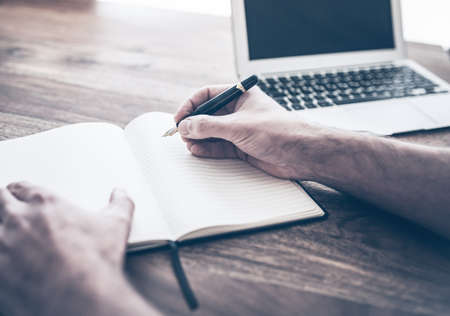 male person writing in notebook besides laptop computer on rustic wooden deskの写真素材