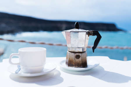 moka pot espresso maker and cup of coffee on table with coastline and ocean in background on sunny dayの写真素材