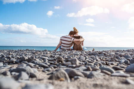 rear view of hugging couple sitting on natural pebble beach against ocean and beautiful skyの写真素材