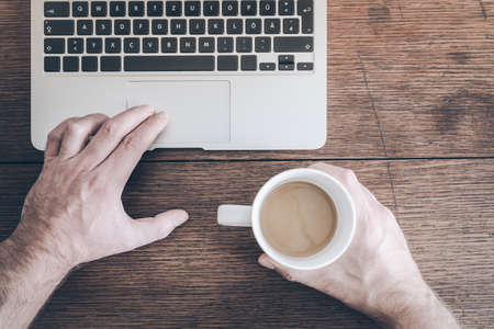 top view of man holding cup of coffee while working on laptop computer on old rustic wooden tableの写真素材
