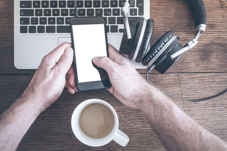 man using smartphone on old wooden table with laptop computer, headphones and cup of coffeeの写真素材
