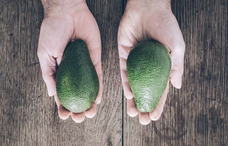 top view of hands of man above old rustic wooden table holding two avocadosの写真素材