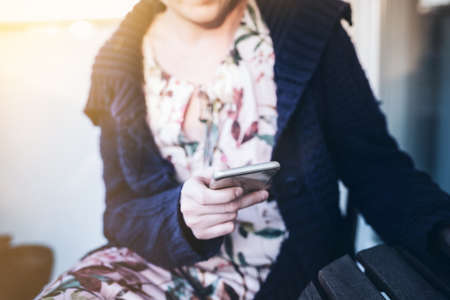 young woman in long dress and cardigan sitting outdoors on porch using her smart phoneの写真素材