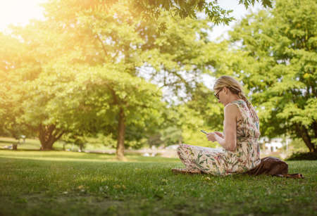 low angle view of woman sitting cross-legged on lawn in public park using her smartphone on sunny summer dayの写真素材