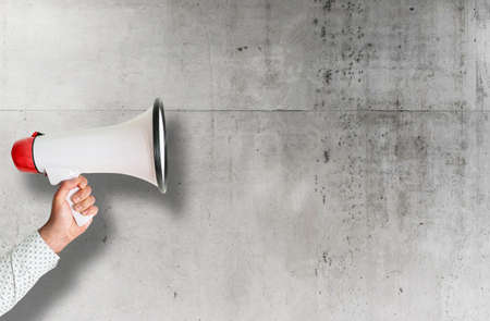 hand holding red and white megaphone against rough concrete wallの写真素材