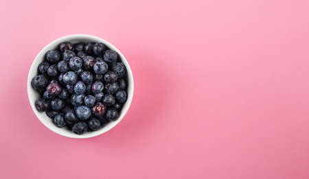 above view of little white pottery bowl filled with fresh ripe blueberries on pink table surfaceの写真素材