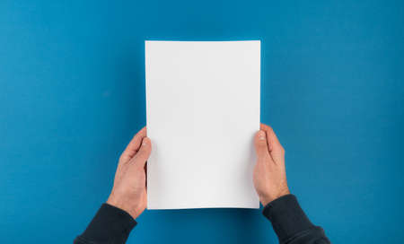 directly above shot of hands holding white blank sheet of paper against blue desk background...