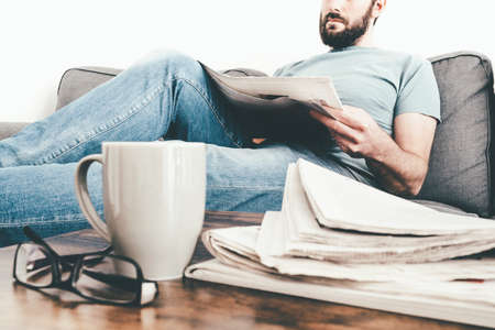 man relaxing on sofa reading a newspaper with stack of newspapers and coffee mug in foreground