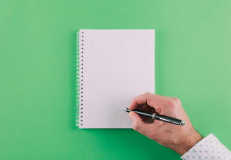 top view of hand holding ballpoint pen against spiral notepad on green background, taking notes...