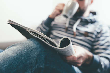 man reading magazine or newspaper and drinking coffee while relaxing on couchの写真素材