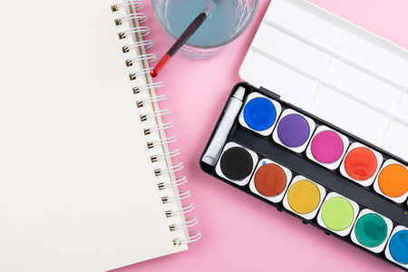 above view of watercolor paintbox, paint brush, sketchbook and glass of water on pink background, painting with watercolors and being creative conceptの写真素材