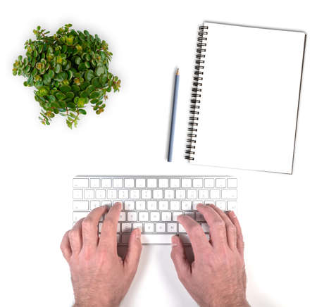 top view of person typing on wireless computer keyboard on white desk with potted plant and notepad with pencilの写真素材