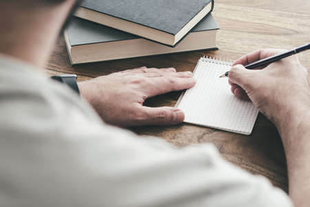 over the shoulder view of man writing on small spiral notepad at wooden table
