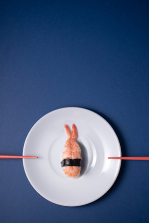 A shrimp sushi on a white plate with pallilos pointing to the center on a blue background.