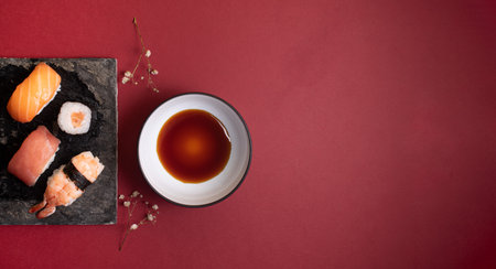 Black plate with four sushi, two sprigs of flowers and a plate of sauce on a red background.の写真素材