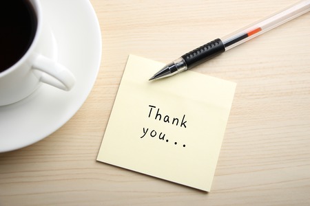 A yellow sticky note is written something on the desk with a cup of coffee and a ball pen aside.の写真素材