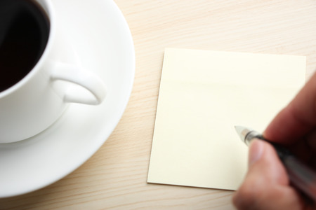 Hand is writing on the blank sticky note with a cup of coffee aside.の写真素材