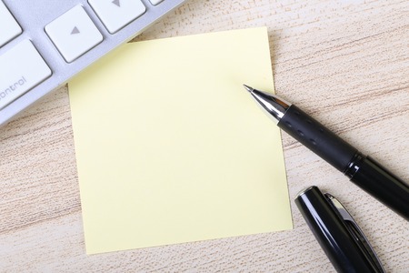 Blank yellow sticky note with keyboard on the wooden desk.