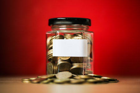 A lot coins in glass money jar with blank white paper on red background.の写真素材