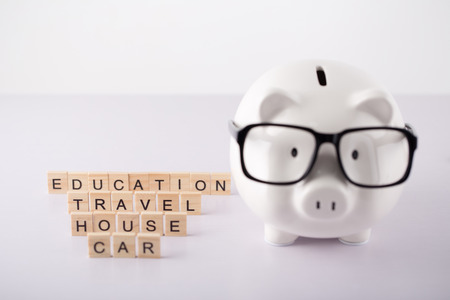 Piggy bank with education, travel, house and car text wood blocks on white table against white background.の写真素材