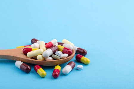 Assorted pharmaceutical medicine pills, tablets and capsules on wooden spoon with blue background.
