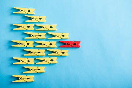 Stand out from the crowd and different concept. Leadership and other similar concepts. One red clothespin stands out from other yellow clothespins on blue background.の写真素材