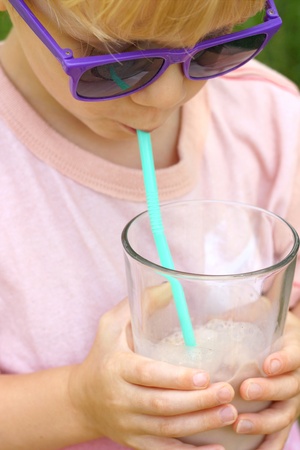 A young child wearing sunglasses is holding a big glass of chocolate milk and sipping it through a straw on a sunny summer dayの写真素材