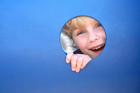 a cute blonde child is smiling and laughing as he peeks his head through a hole in a playground tunnel on a summer dayの写真素材