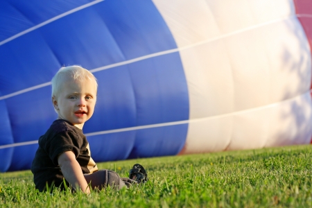 a cute baby boy is sitting in the grass on a summer day, watching a red, white, and blue hot air balloon fill up.の写真素材