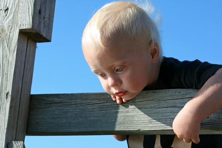 a close up on cute baby who is standing on a wooden fence outside on a summer day, hanging over the edge and looking down into the water below.の写真素材