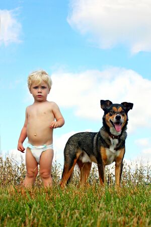 a cute little blonde baby boy is standing outside on a beautiful summer day with his pet German Shepherd Dog, with the blue sky behind them.の写真素材