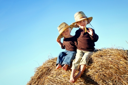 Two children, a young boy and his baby brother are sitting outside on hay bales, wearing straw hats on a sunny Autumn day  Room for Text, Copy Space の写真素材