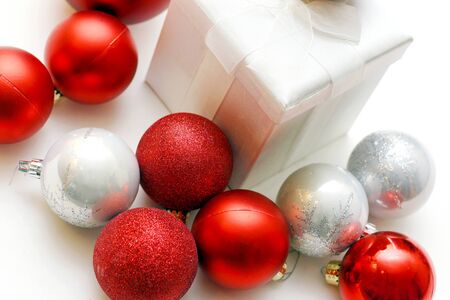 a white christmas present with a bow is sitting in front of an isolated white background, with silver and red sparkling bulb ornaments around it.の写真素材