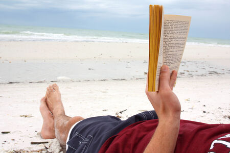 the hands and legs of a relaxing man can be seen laying on the beach by the ocean, reading a bookの写真素材