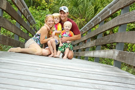 a young, happy family of four people including mother, father, baby and child are sitting on a wooden bridge in front of a tropical forestの写真素材