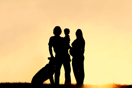 A silhouette of a happy family of three people, father, child, and pregnant mother, and their dog outside in front of the sunset の写真素材