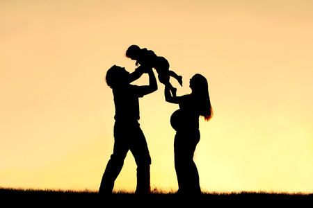 A silhouette of a happy family of three people, father, child and pregnant mother, are celebrating and praising God outside at sunset の写真素材