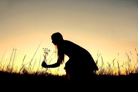 A silhouette of a happy girl outside picking wild flowers in the meadow at sunset の写真素材