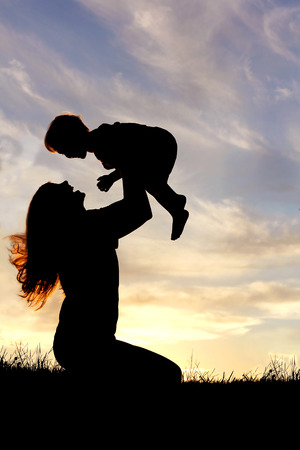 A silhouette of a happy young mother, laughing as she plays with her toddler child and lifts him over her head outside at sunset.の写真素材