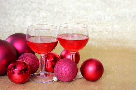 Two wine glasses fill with red beverage are sitting in a toast surrounded by Christmas tree bulb decorations in front of a glittery gold .の写真素材