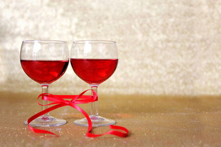 Two glasses of red wine are tied together with a red ribbon bow in the form of a Christmas or New years Eve toast, in front of a sparkling gold .の写真素材