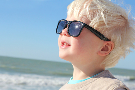 a happy young child wearing sunglasses is smiling as he basks in the summer sun on the beach by the ocean.の写真素材