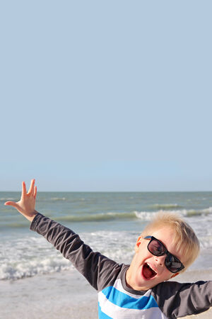 A super happy young child is smiling with joy with his arms raised up while standing on the white sand beach by the ocean on summer vacation.の写真素材