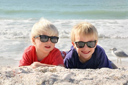 Two happy young children, a little boy and his toddler brother, are laying on the white sand beach in the sun, wearing sunglasses on family summer vacation.の写真素材