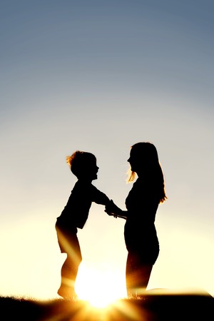 Silhouette of a young mother lovingly holding hands with her happy little child outside in front of...