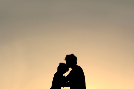 A silhouette of a Father lovingly kissing his young child on the head at sunset.の写真素材