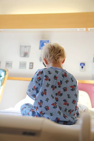 The back of a young boy child can be seen sitting in the bed at a bright hospital.の写真素材