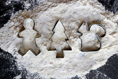 A collection of silver Winter decoration Christmas Cookie cutters in a pile of white baking flour on a black slate background.の写真素材