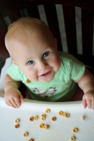 A happy 8 month old baby girl is smiling at the camera as she is sitting in her high chair eating hold grain finger food cereal.の写真素材