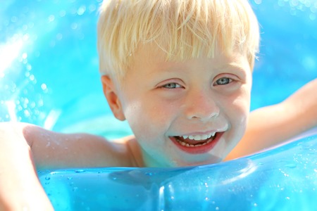 A cute, blonde, three year old boy child is laughing as he floats on a blue inner tube in the pool on a sunny summer day.の写真素材